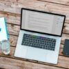 silver MacBook beside space gray iPhone 6 and clear drinking glass on brown wooden top